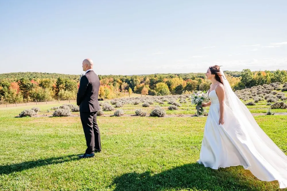 The bride walks up behind the groom across the lawn for their first look