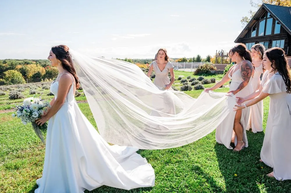The bride's long veil blows sideways as bridesmaids laugh behind her on the lawn