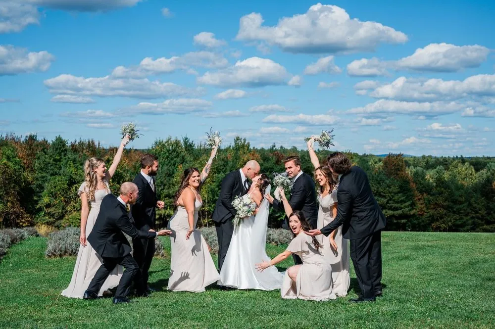 The wedding party cheers and hoists bouquets on a hilltop lawn under puffy clouds