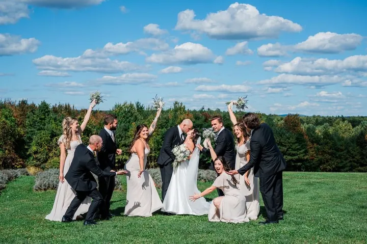 The wedding party cheers and hoists bouquets on a hilltop lawn under puffy clouds