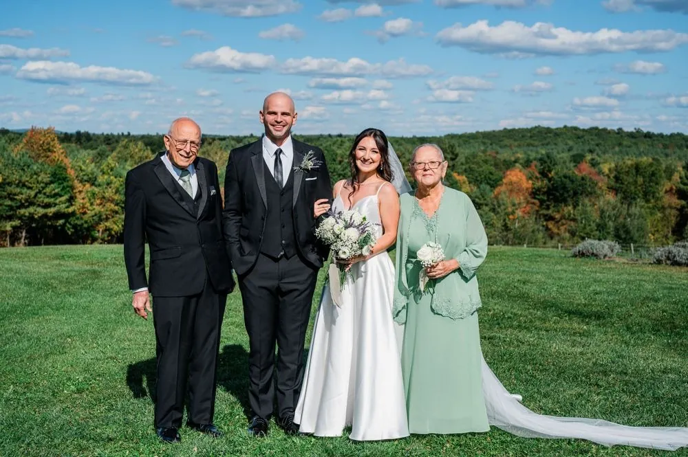 The couple poses with 2 older guests on the lawn, trees changing color across the valley