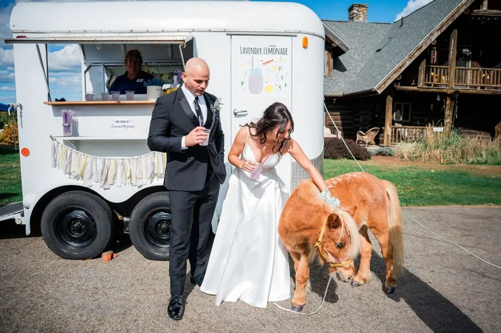The bride pets a chestnut pony beside a white food trailer while the groom watches