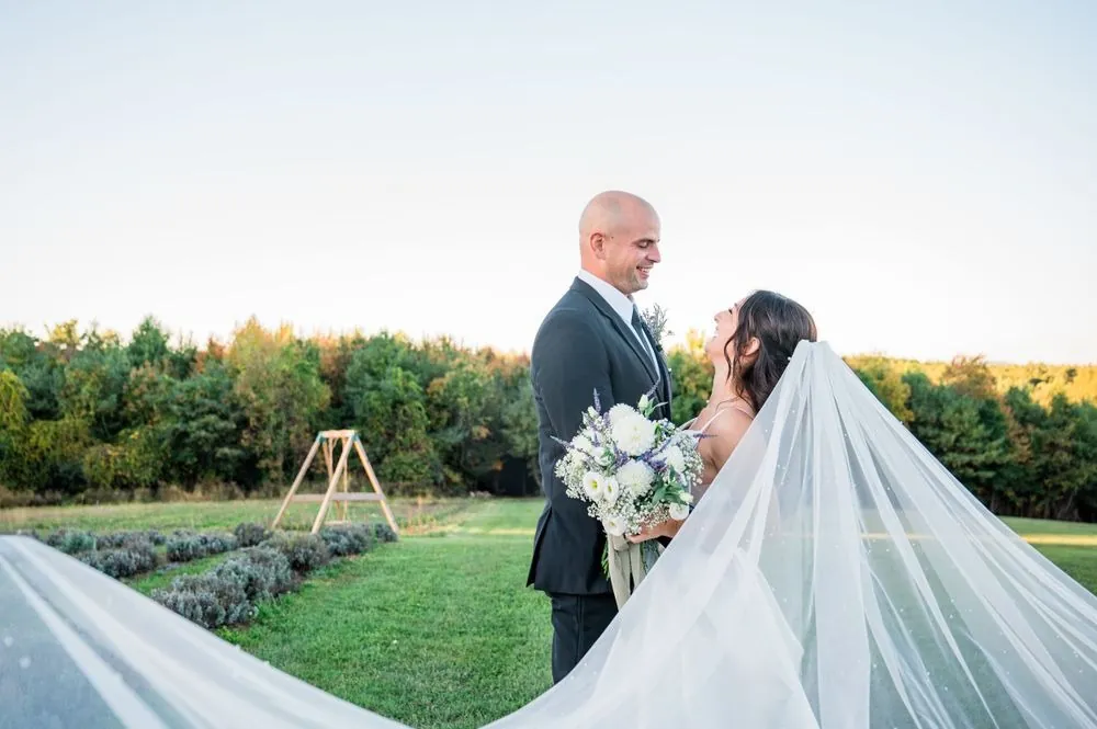 Kailey and Patrick smile at each other as her long veil sweeps across the lawn