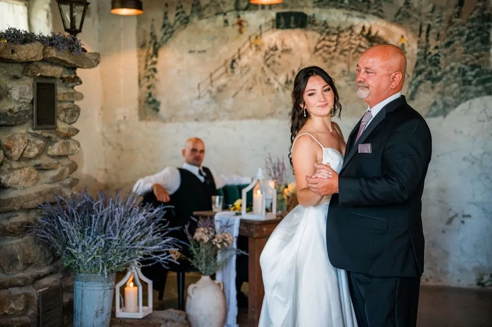 The bride slow dances with an older man in a dark suit beside dried lavender and lanterns