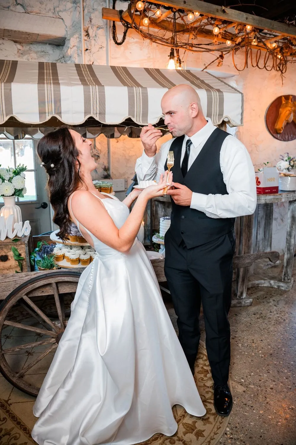The groom takes a bite of cake as the bride laughs under a striped awning cart