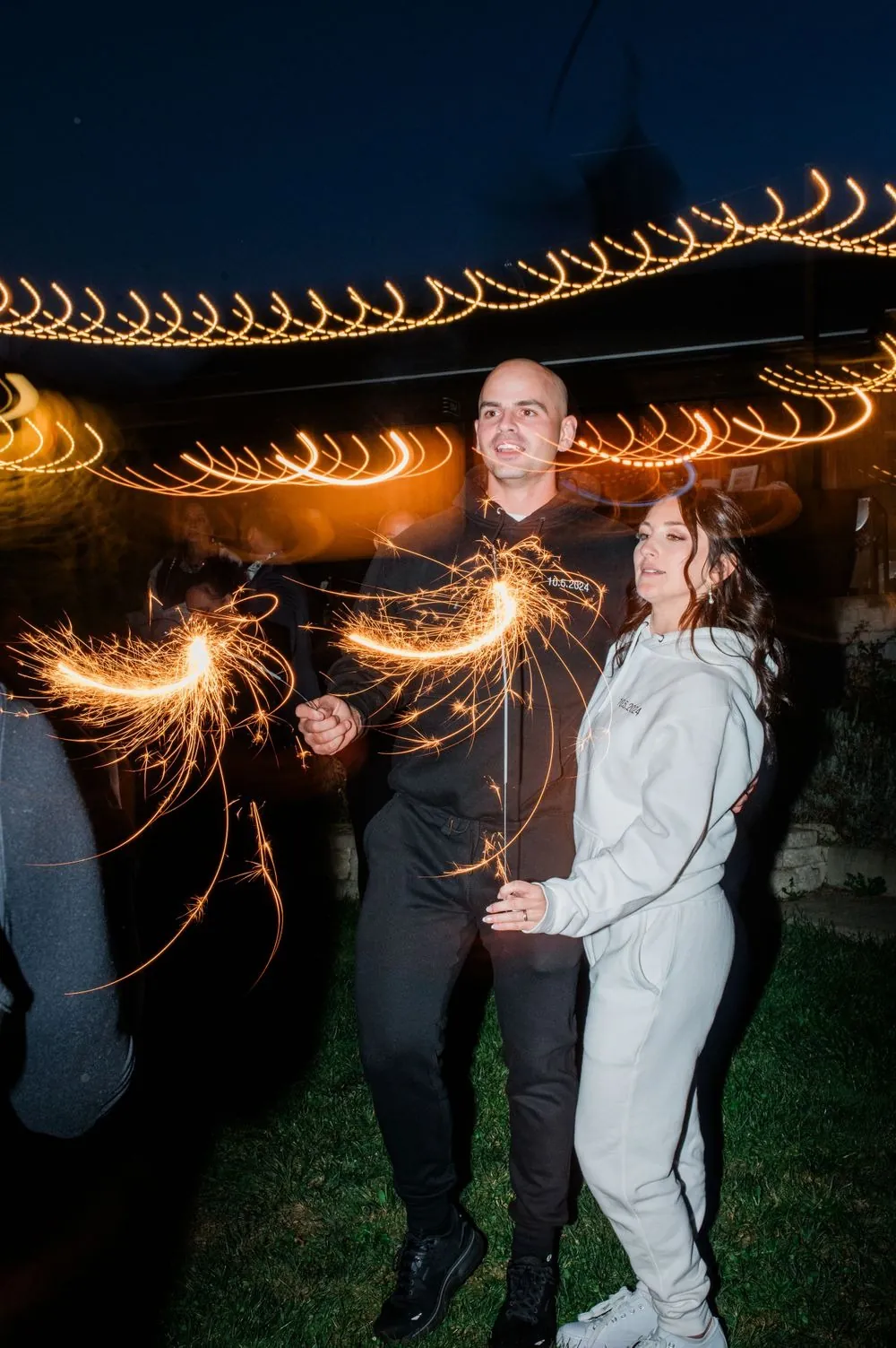 Kailey and Patrick in matching sweatsuits wave sparklers under string lights at night