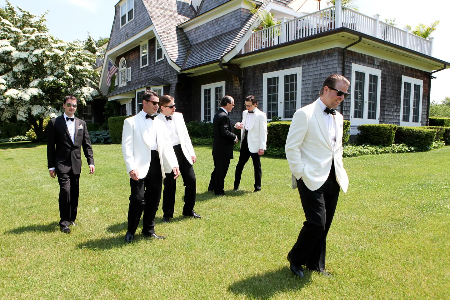 Groomsmen in white dinner jackets and sunglasses stroll across the lawn of a shingled house