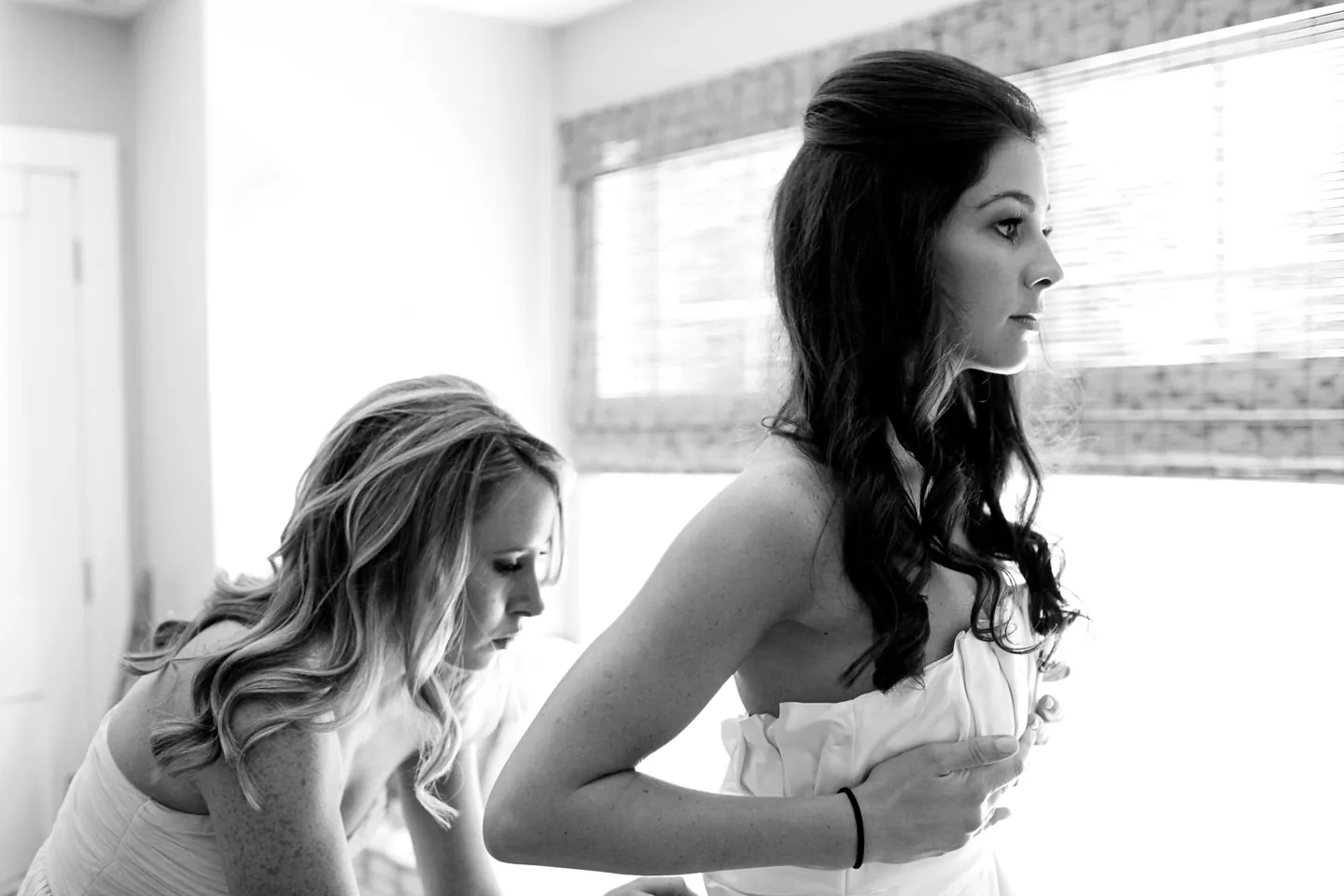 Black and white frame of the bride fixing her strapless gown as a friend helps beside a window