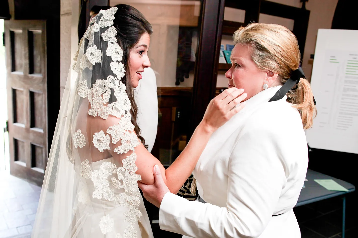 A woman in a white jacket fusses over the bride, who wears a long lace-trimmed veil by a dark door