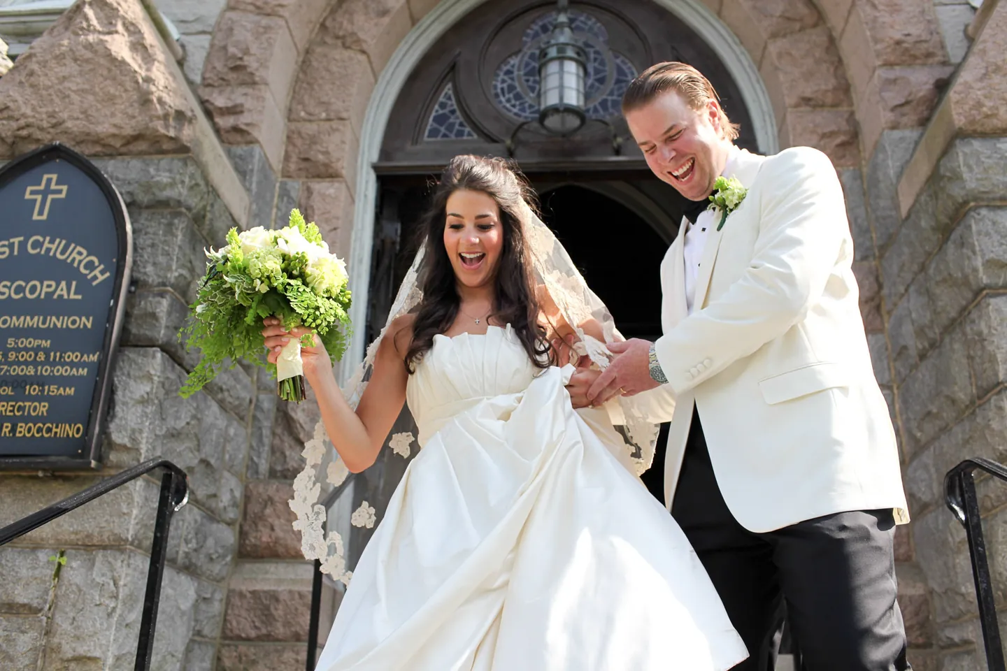 Kaitlin and Matthew step out of the stone church doorway, her green and white bouquet raised