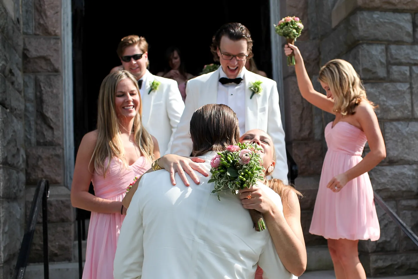 Bridesmaids in pale pink hug the couple on the church steps, one waving her bouquet overhead