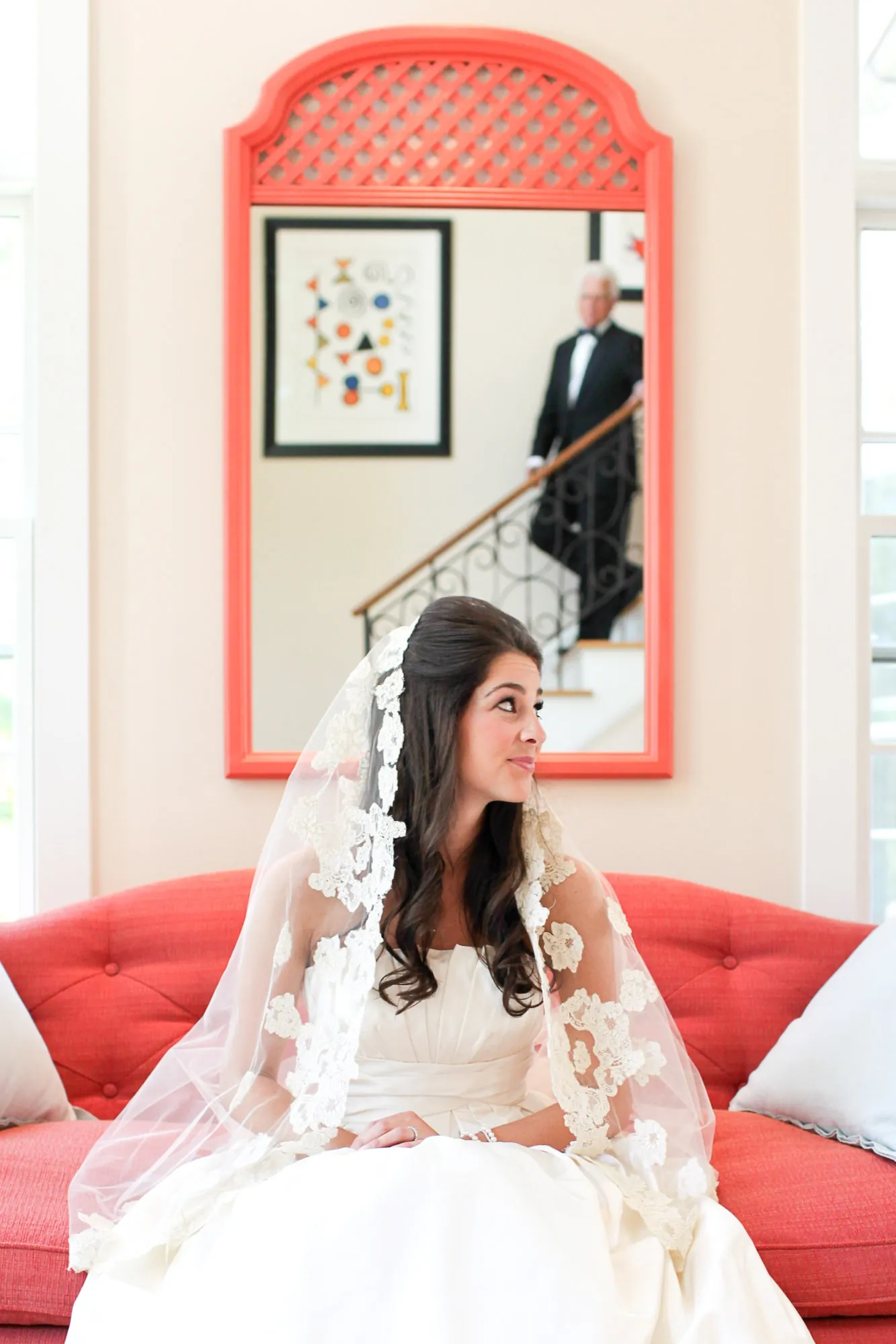 The bride sits on a red sofa under a coral-framed mirror, wrapped in a lace-edged veil