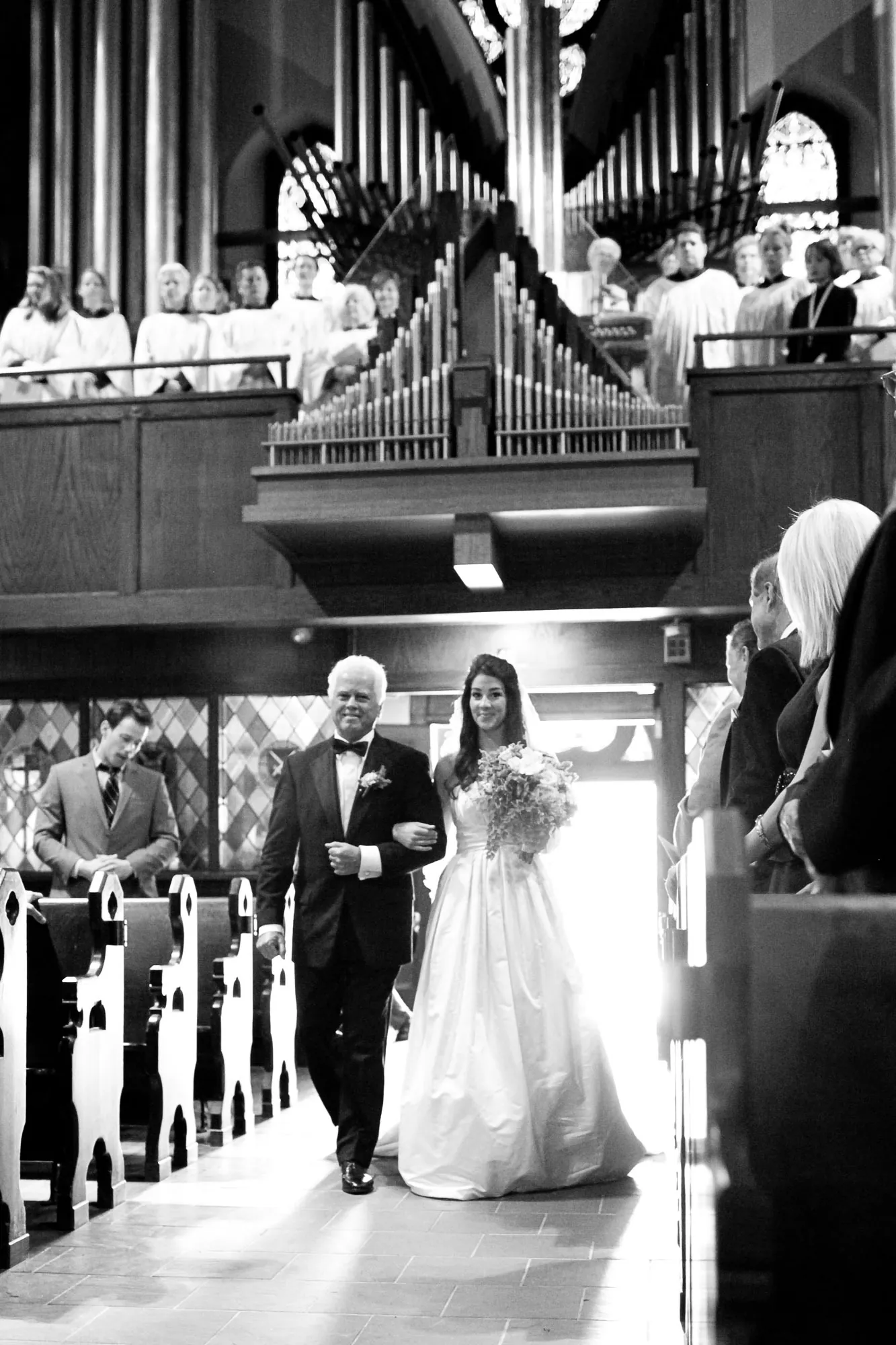 Black and white frame of the bride walking the aisle with an older man beneath organ pipes