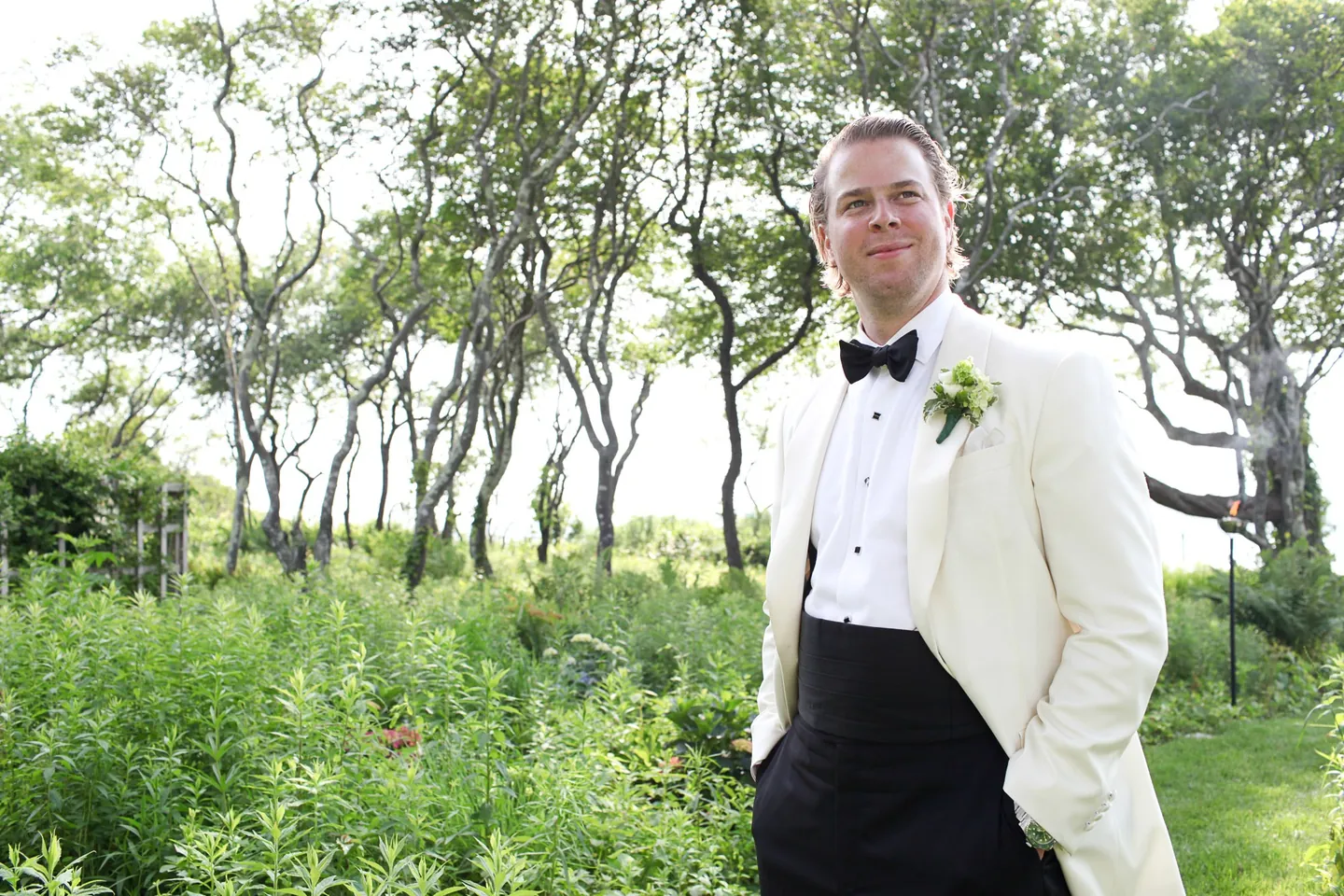 The groom in a white dinner jacket stands among wind-shaped shrubs and trees