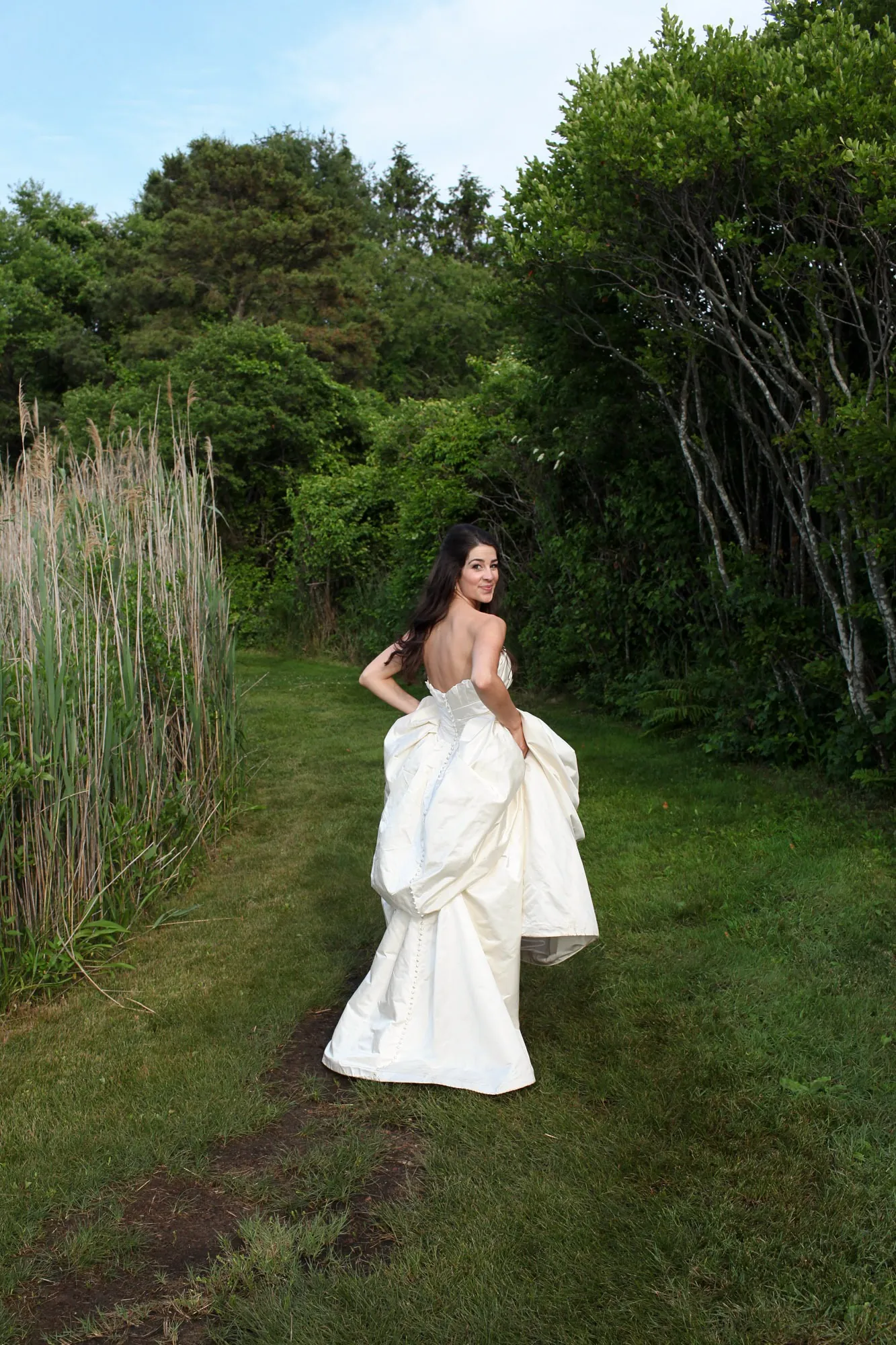 The bride walks alone down a mown path through tall green grasses