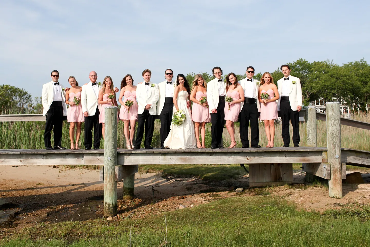 The wedding party in white jackets and pink dresses lines a wooden boardwalk over the marsh