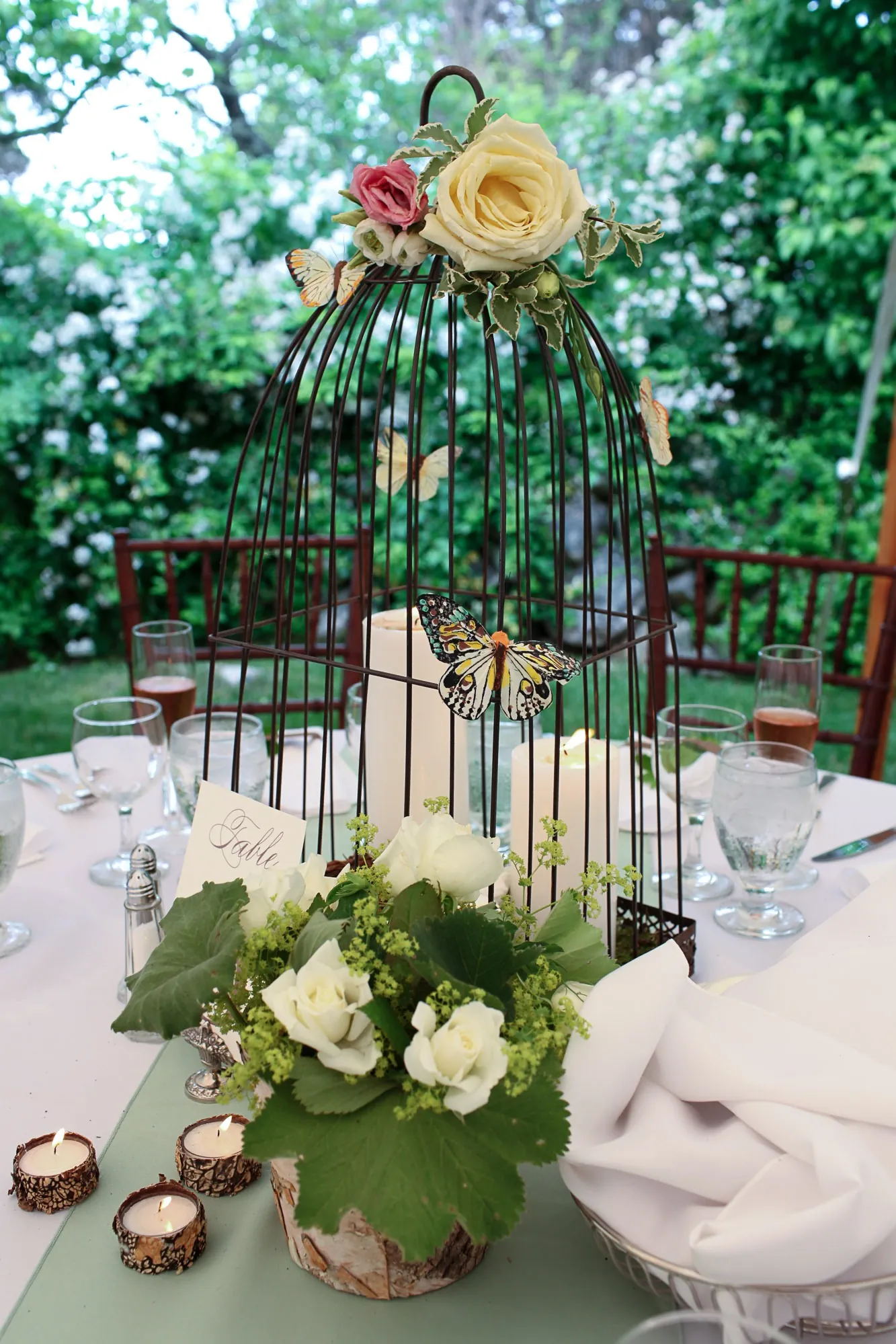 A black wire birdcage topped with roses and butterflies above white flowers on a reception table