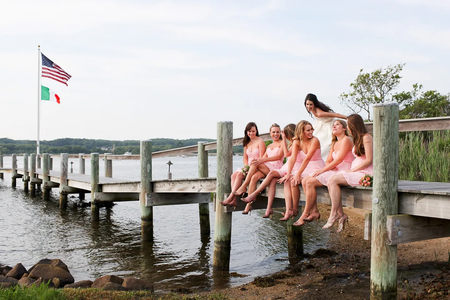 Bridesmaids in pink sit in a row along the dock, legs dangling over the water