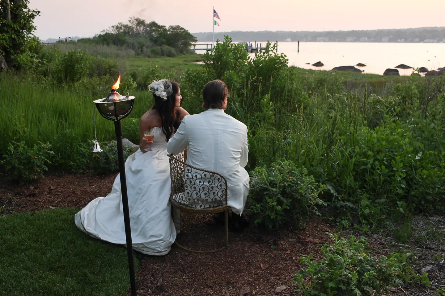 The couple sit with drinks beside a lit tiki torch, looking out over the bay