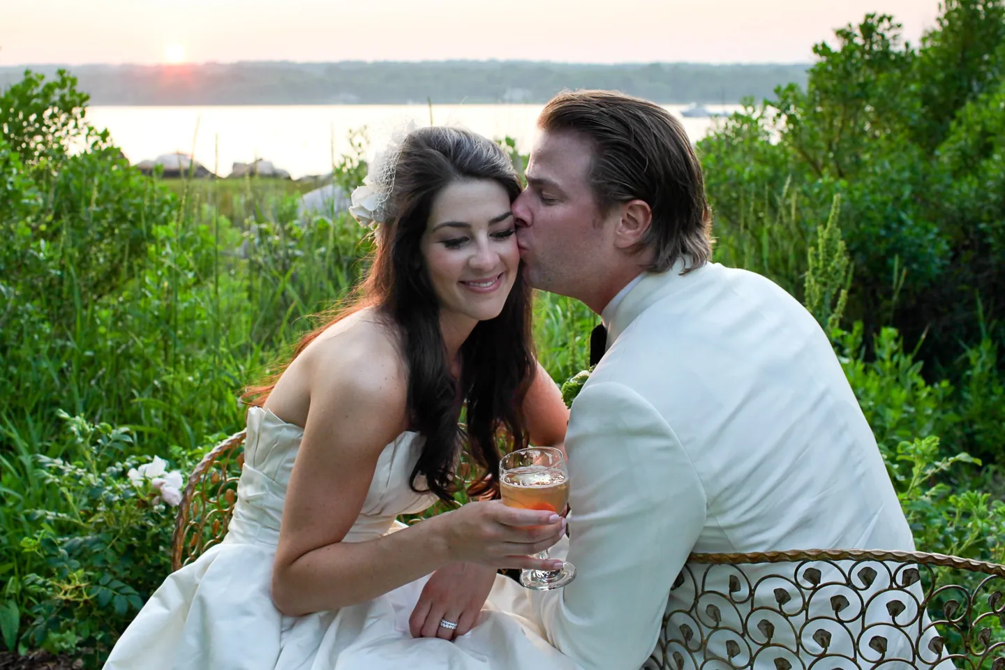 Matthew kisses Kaitlin's cheek on a curled metal bench as the sun sets over the water