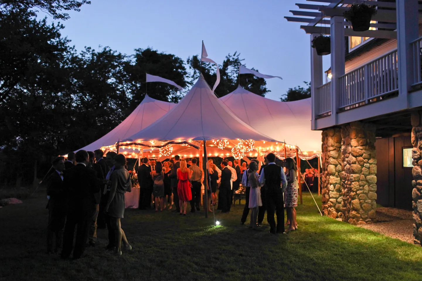 Sailcloth tents glow warm at dusk as guests mingle on the lawn beside the house