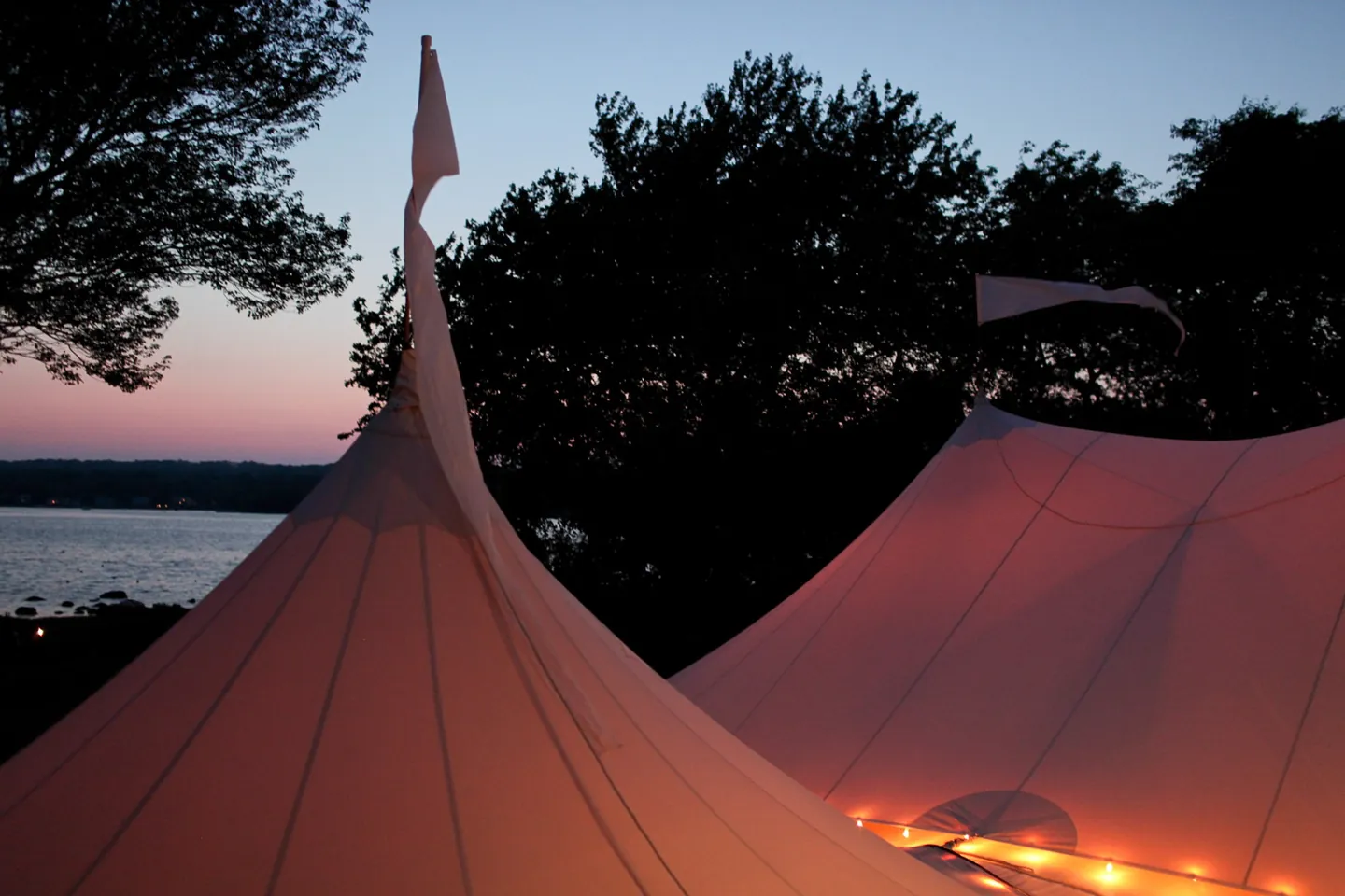 Peach-lit tent peaks silhouetted against dark trees with the bay behind at twilight