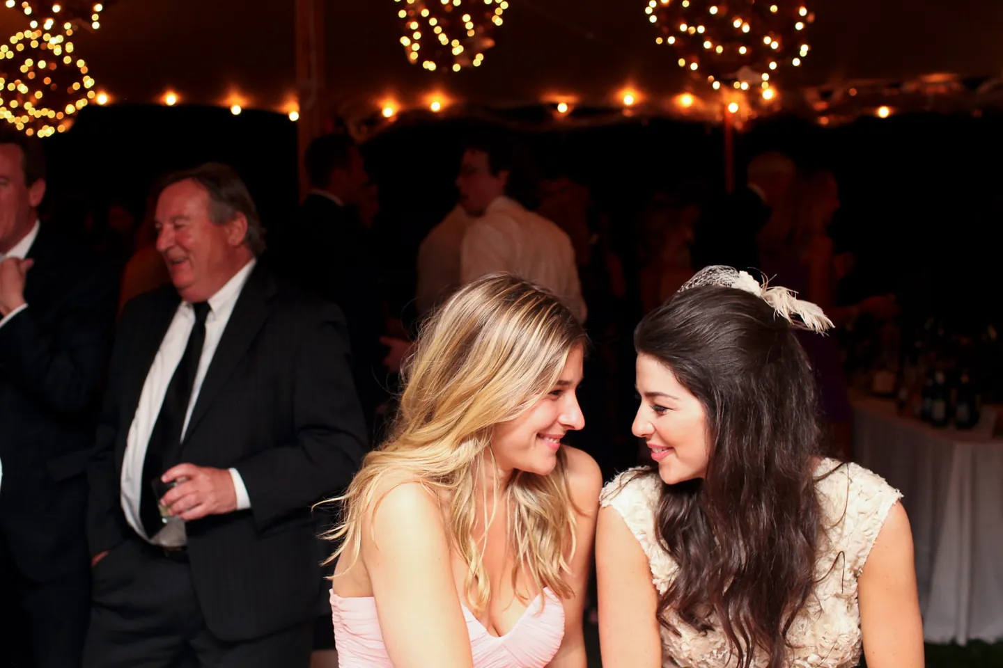 The bride and a friend in pink lean close and laugh beneath glowing lantern spheres