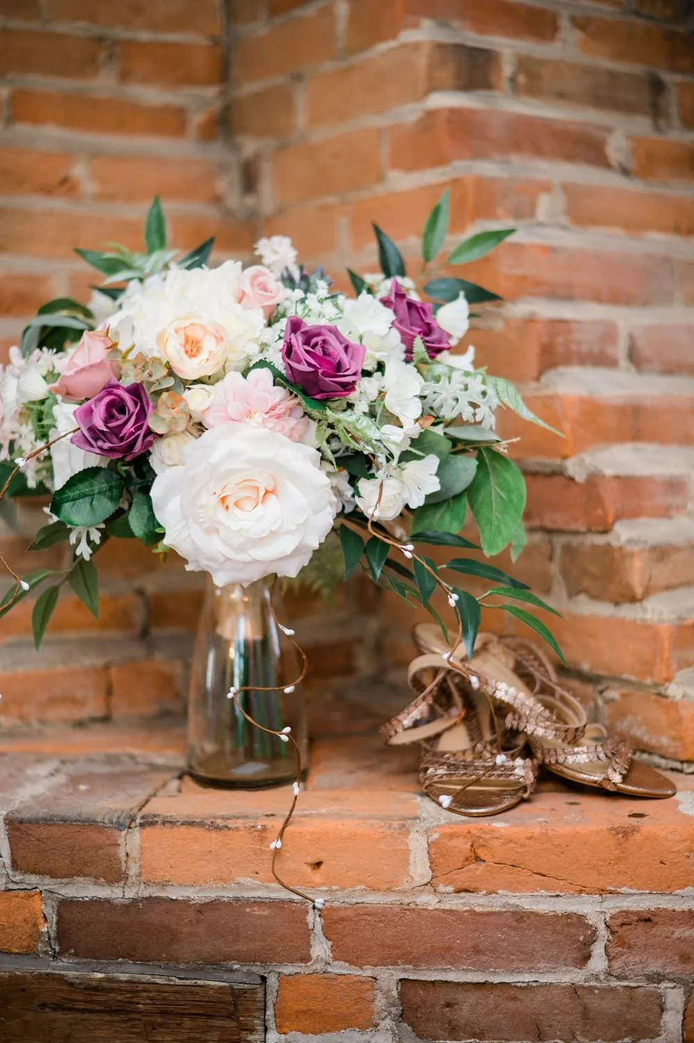 The bridal bouquet of blush roses and greenery rests in a vase against a brick wall