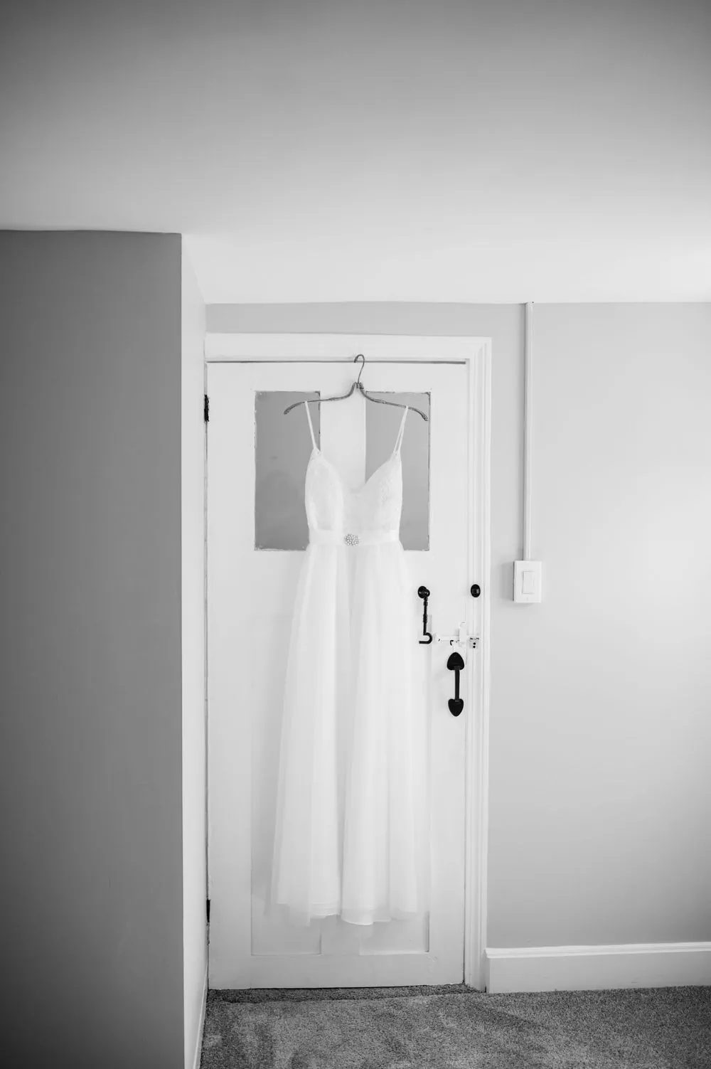 The wedding gown hangs on a white door in soft window light, in black and white