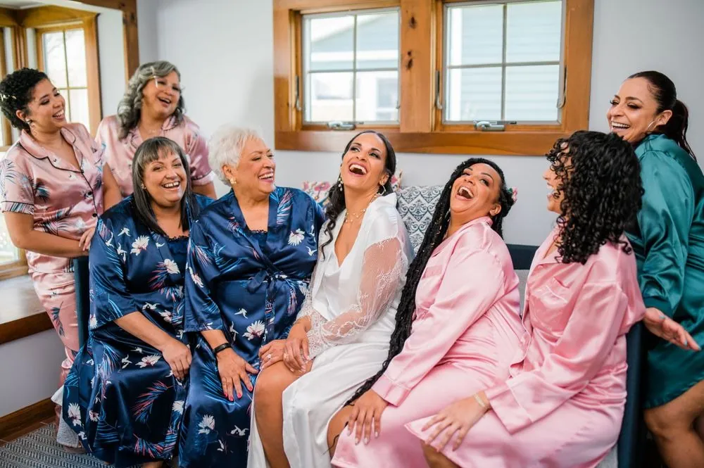 The bride in a white robe laughs with women in pink and navy satin robes on a sofa