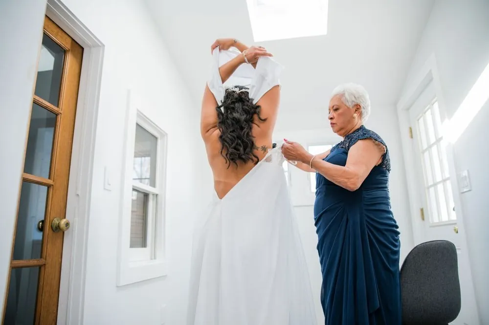 An older woman in navy fastens the back of the bride's gown by a sunny window