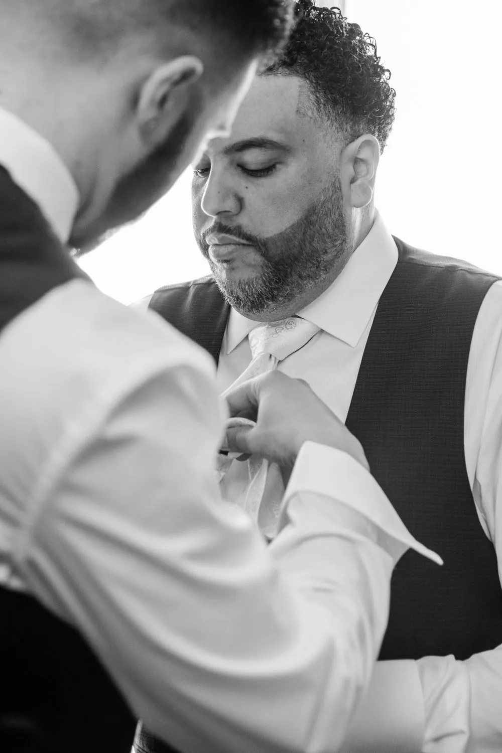 A groomsman adjusts the groom's tie as he looks down, in black and white