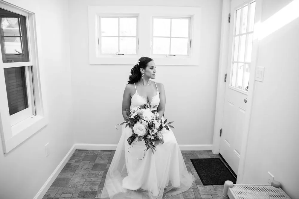 The bride stands in a bright white hallway holding her bouquet, in black and white