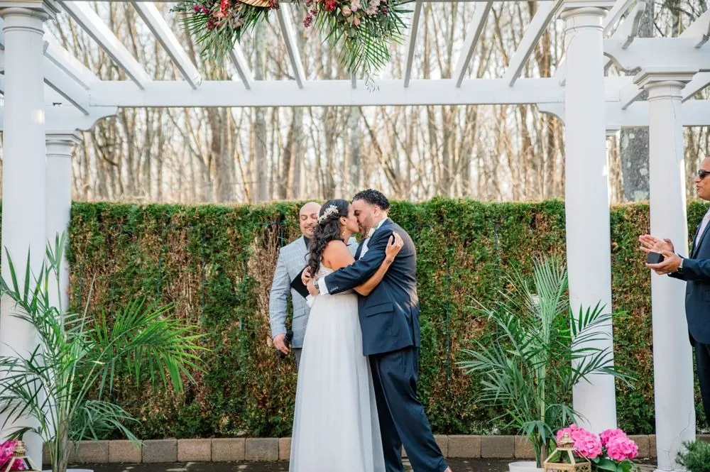 Karla and Carlos kiss under the white pergola as bridesmaids in dusty rose watch