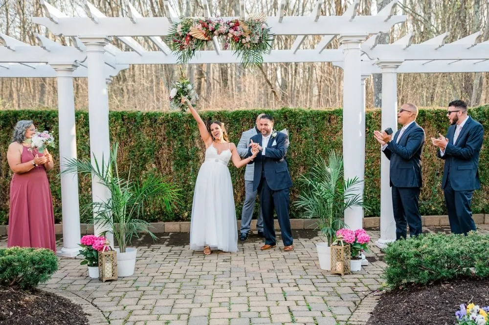 The wedding party cheers and raises bouquets around the couple under the pergola