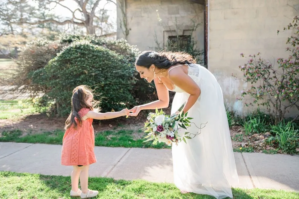 A little girl in a coral dress holds the bride's hand on a sunny garden path