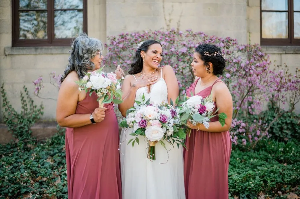 The bride laughs with 2 bridesmaids in rose gowns in front of purple azaleas