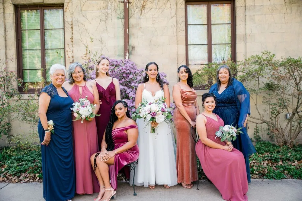 Bridesmaids in rose, mauve and navy gowns pose with the bride against a stone wall