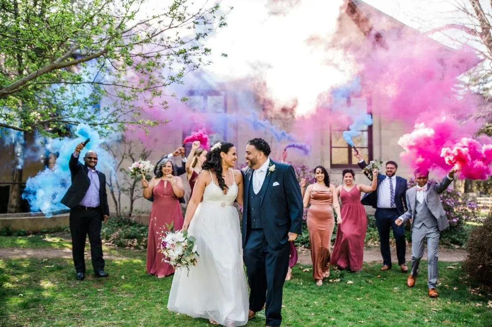 Karla and Carlos walk arm in arm as the wedding party waves pink and blue smoke behind them