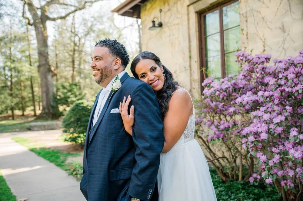 Karla rests her head on Carlos's shoulder beside a purple azalea in full bloom
