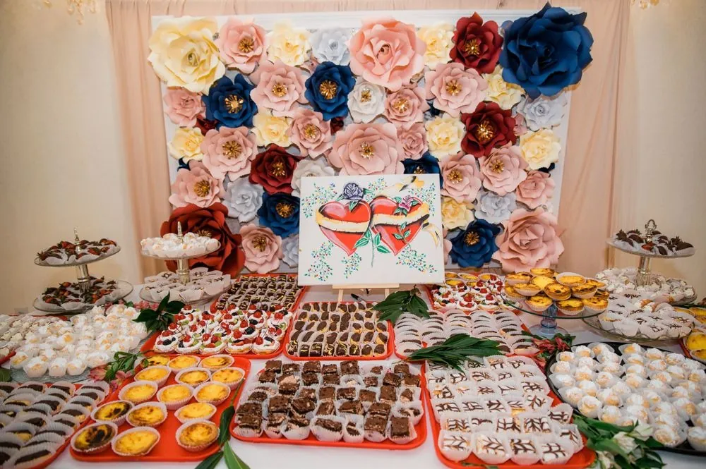 A paper-flower wall rises over a dessert table covered in trays of pastries and sweets