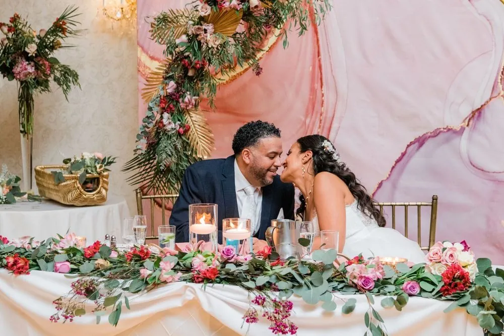 Karla and Carlos kiss at the sweetheart table behind a garland of pink and red roses