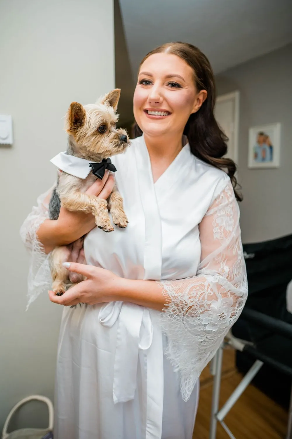The bride in a white lace robe smiles while holding a small Yorkie