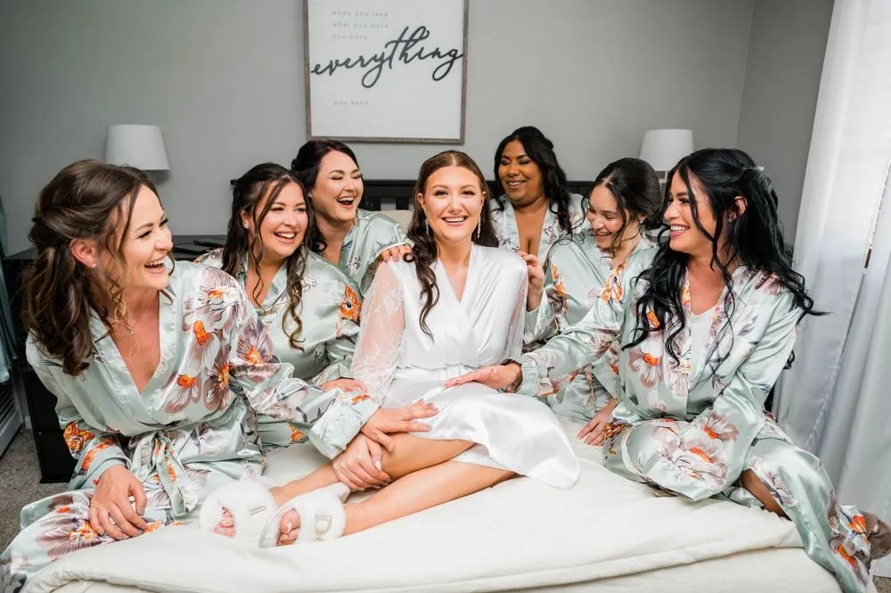 Bridesmaids in sage floral robes crowd around the bride on a white bed