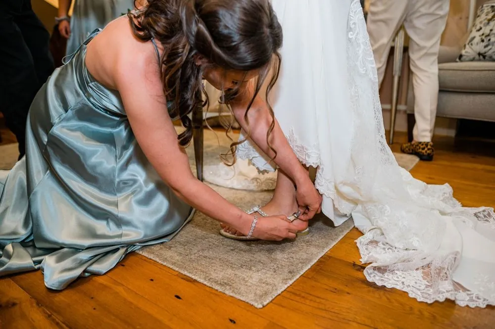 A bridesmaid in sage kneels to buckle the bride's shoe beside her gown