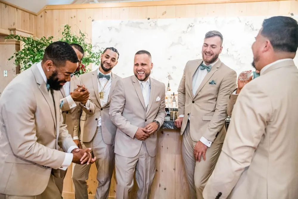 Groomsmen in sand-colored suits laugh together in a bright room