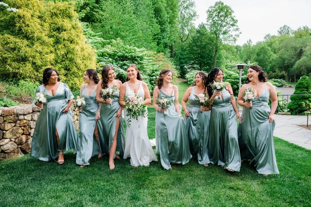 Bridesmaids in sage satin gowns laugh together on the green lawn