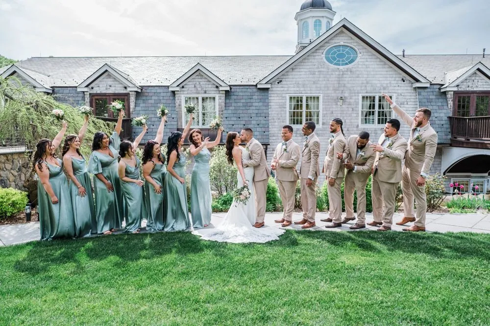 Wedding party cheers and raises bouquets as Kasey and Derek kiss before the shingled clubhouse