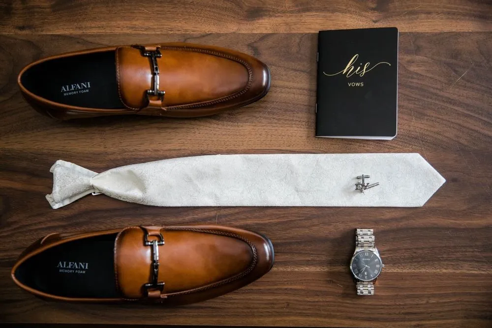 Cognac loafers, a silver tie, a watch and a black vow book on dark wood