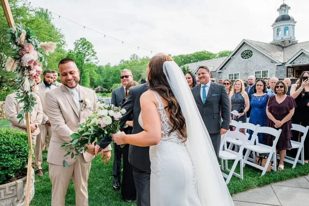 The groom grins as the bride's hand is placed in his at the floral arch